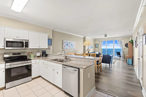 Open-plan kitchen and living room with white cabinetry, granite countertops, stainless steel appliances, and ocean view through glass doors.
