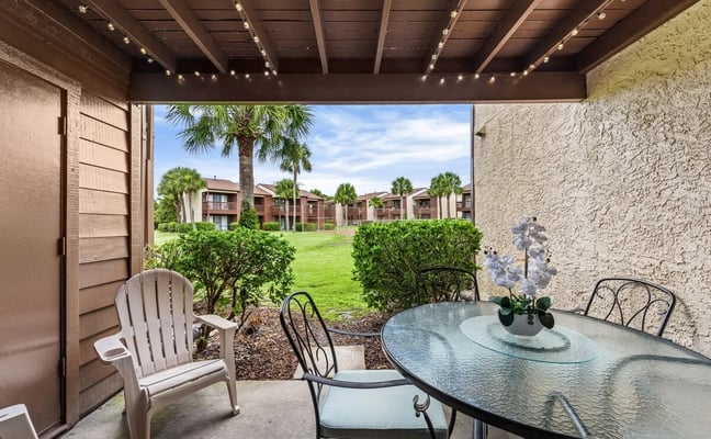 Modern two-story beach house with white siding, covered front porch with turquoise Adirondack chairs, upper deck, and palm trees in sandy yard.